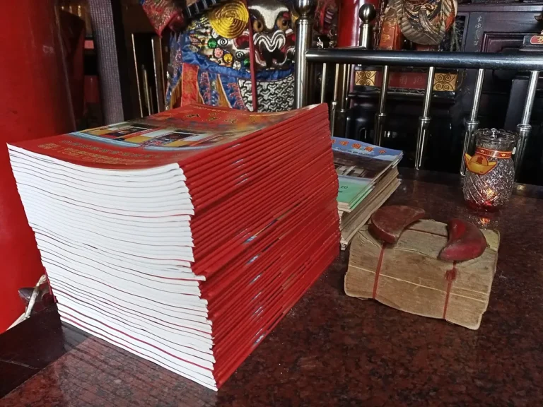 Free lunar-calendar almanac booklets stacked on a table at Jindun Chenghuang Temple, Taiwan, beside jiaobei divination blocks and joss paper