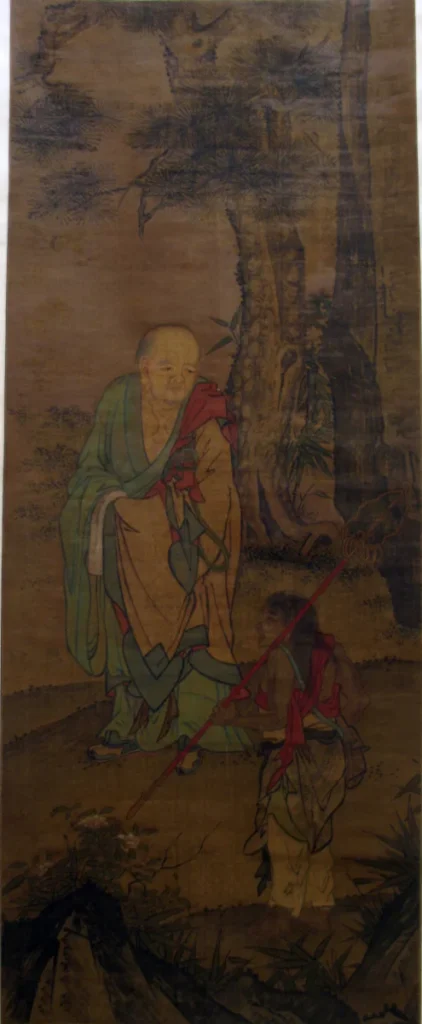 A luohan in red and green robes standing beneath rocks and trees on a hanging scroll, with a small attendant figure at the lower left; ink and colour on silk.